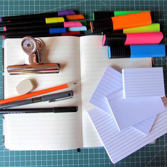 Open notebook on a grid mat with stationary supplies including, colorful pens, highlighters, a clip, eraser, pencil, pen, and index cards.