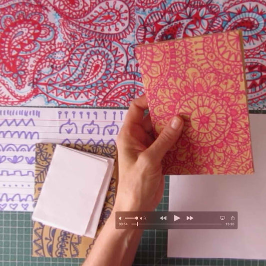 A person holds a pink mandala doodle on a piece of paper over a work desk with more examples, paper,  and a chart with shapes. 