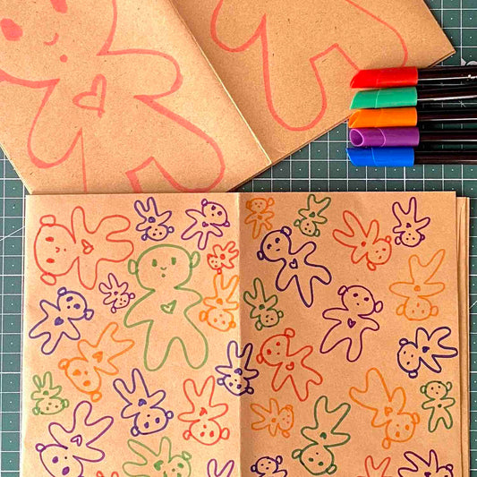 Two brown papers decorated on the front with doodles of colorful dolls in different sizes by Alex Mitchell, with markers on a green work surface.