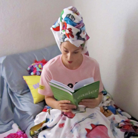 Alex Mitchell relaxing reading green book with a colorful cozy throw blanket and wearing a fun beach towel on her head.