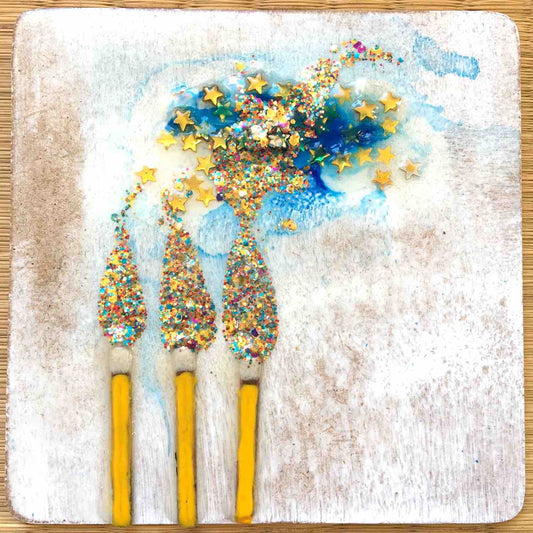 Three matchsticks with colorful glitter and gold star confetti resembling flames and smoke on white background by artist Alex Mitchell.
