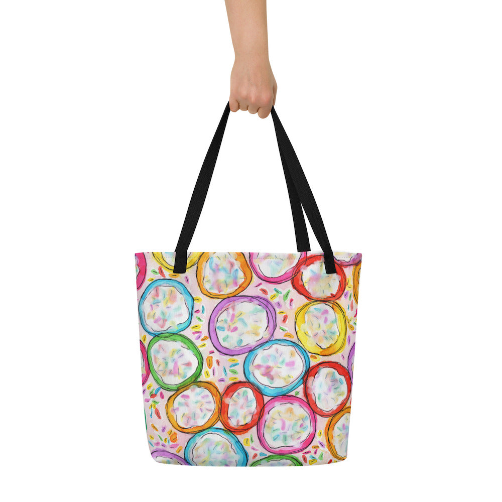 Big sturdy graphic tote bag with multicolor Frosted Cookies print on front and black straps held by hand.