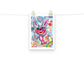 Colorful happy fine art print of a pink rabbit waving hello on vibrant background hanging on wall from a string with gold clips.