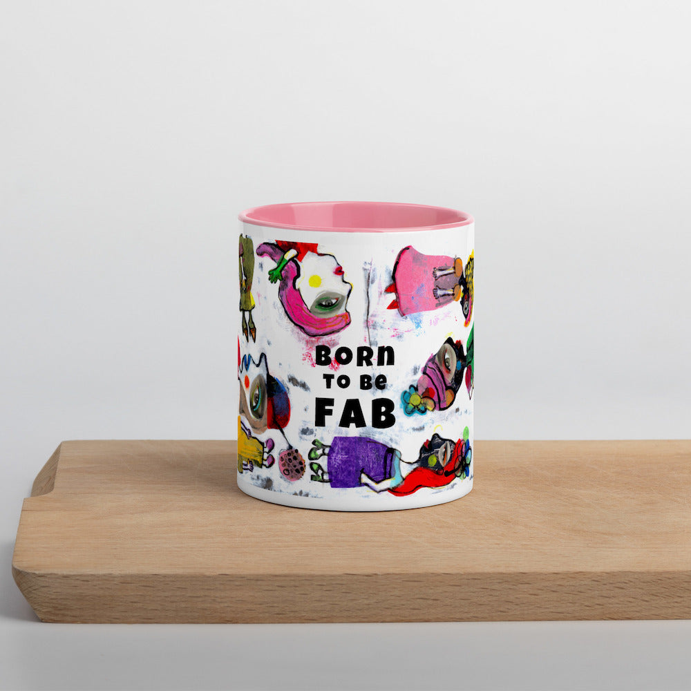 Graphic coffee mug with slogan Born To Be Fab and pink accent color on rim and interior on wood counter front view.